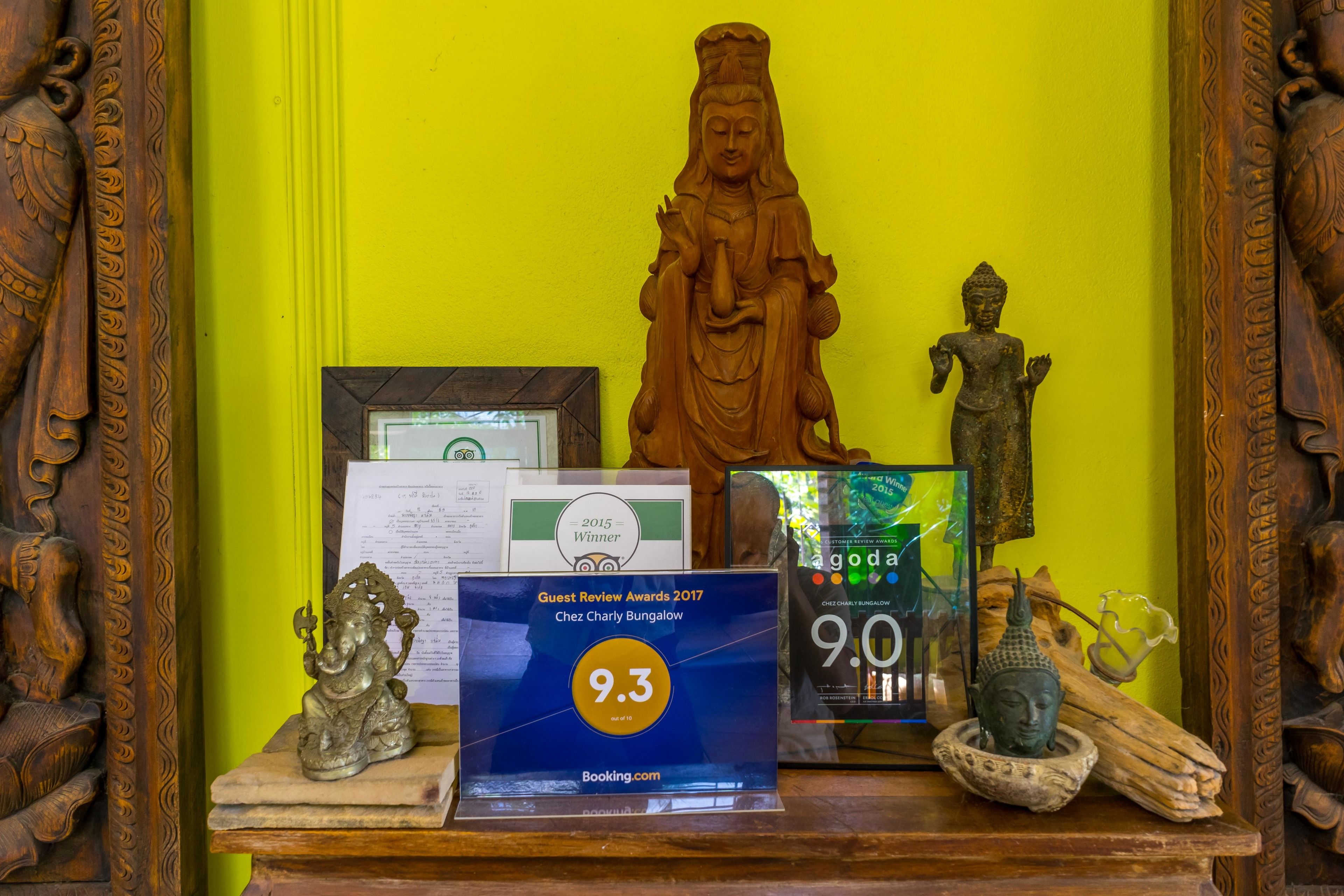 Una vitrina de madera con una estatua de Buda, un certificado enmarcado, una placa de reconocimiento por reseñas de huéspedes y una pequeña figura de Buda sobre una mesa.