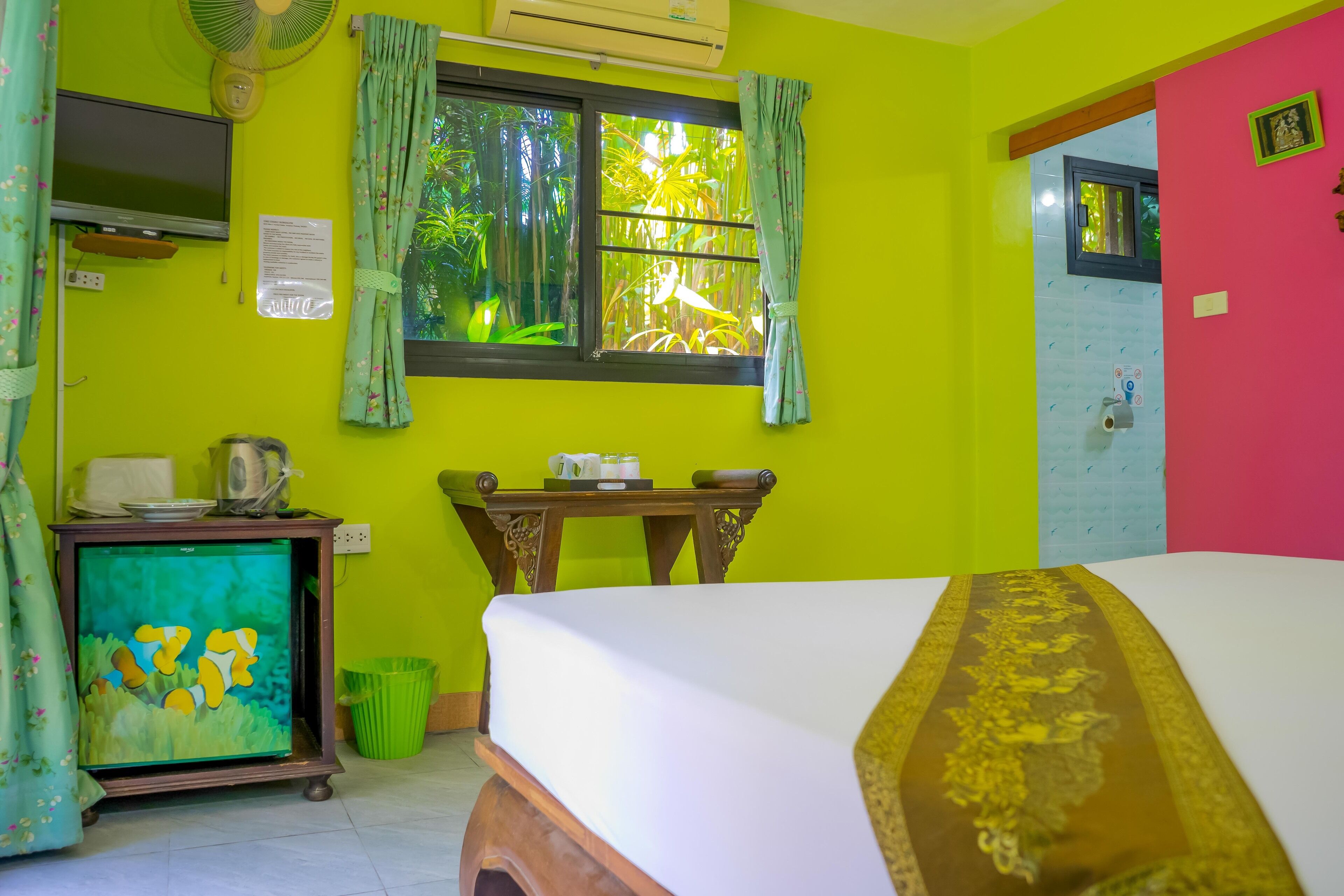 A room with a bed, a desk with a computer, a window with curtains, and a fan.