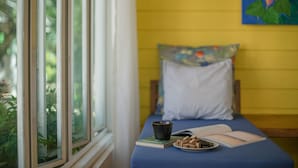 A room with a bed against a yellow wall, a window with greenery outside, and a small table with a book, a cup, and some snacks. - Lemon House (Patong)