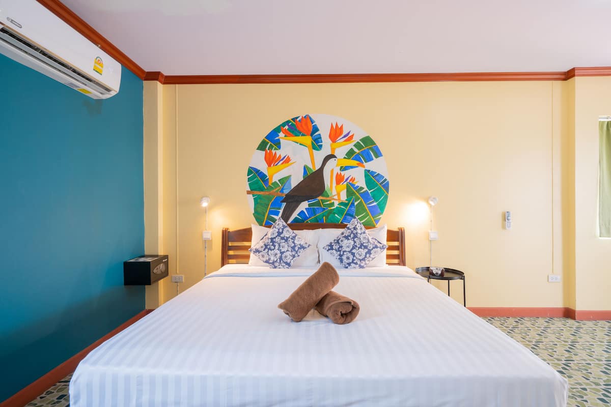 A hotel room with a bed, a wall-mounted air conditioner, a decorative wall piece, and a nightstand with a lamp.