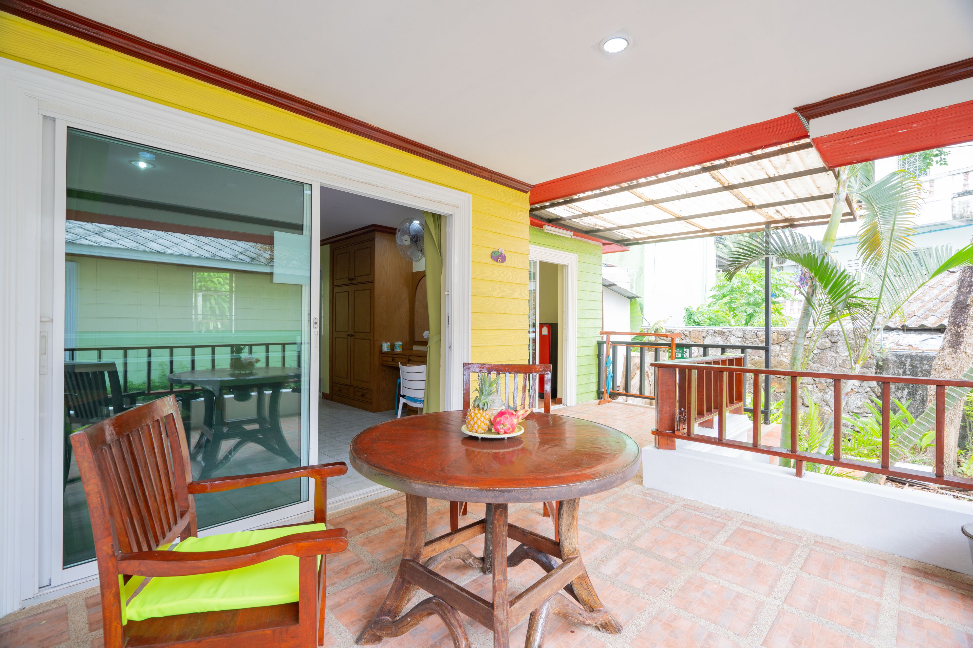 A covered outdoor area with a wooden table and chairs, a sliding glass door leading to a balcony, and a view of a garden.