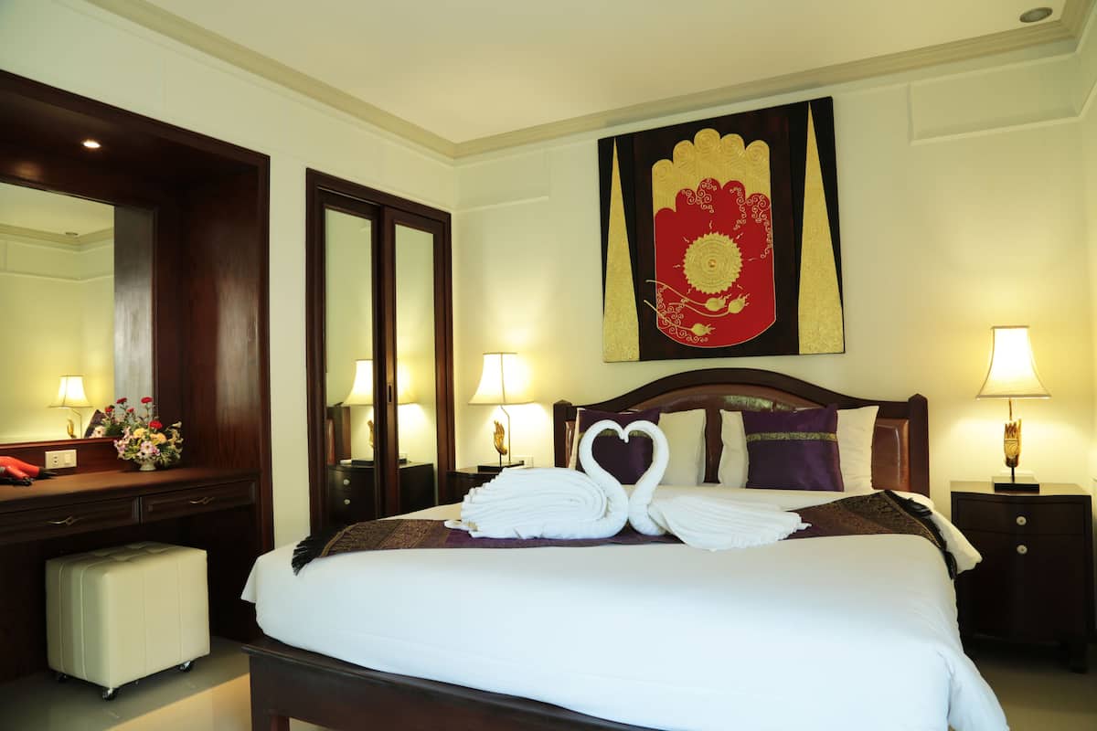 A hotel room with a neatly made bed, a large mirror, a decorative wall hanging, and a desk with a lamp.