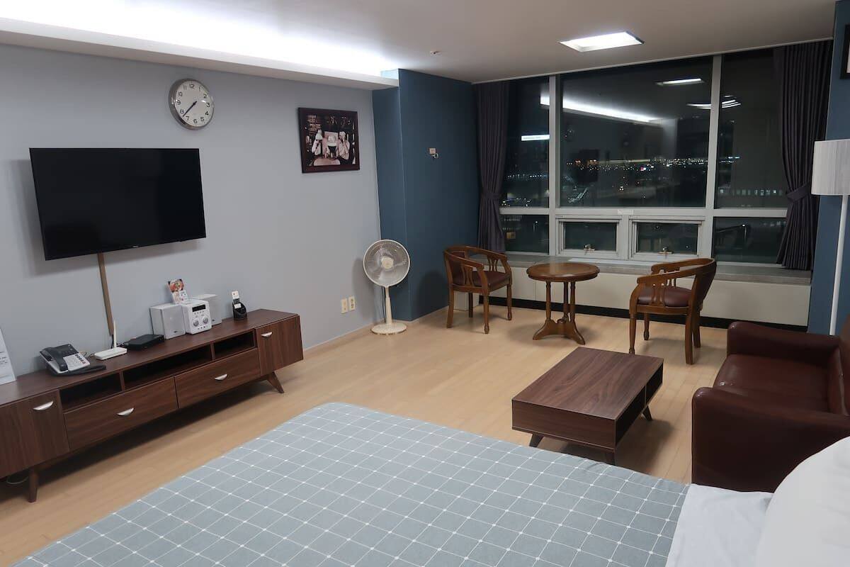 A hotel room with a bed, a TV, a desk, a chair, a fan, and a window with a city view.