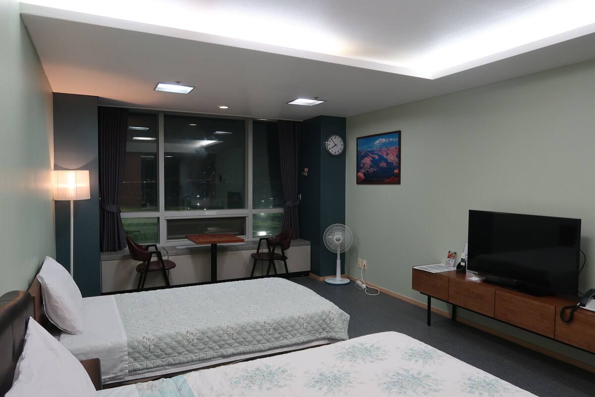 A hotel room with two beds, a desk with a chair, a TV, a fan, and a window with curtains.