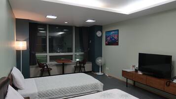 A hotel room with two beds, a desk with a chair, a TV, a fan, and a window with curtains.