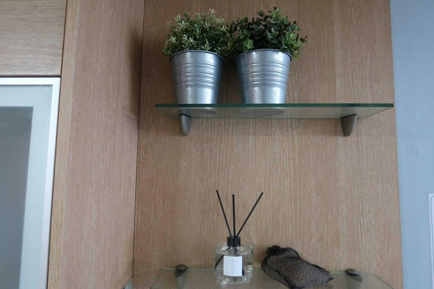 A wooden wall with a glass shelf holding two potted plants and a glass table with a bottle and a pair of slippers.