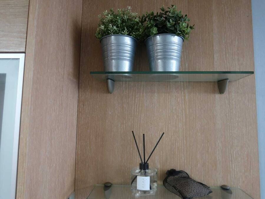 A wooden wall with a glass shelf holding two potted plants and a glass table with a bottle and a pair of slippers.