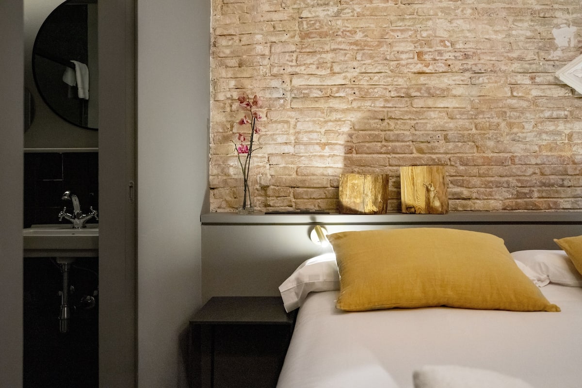 Un dormitorio con pared de ladrillo, cama con ropa blanca y almohada color mostaza, mesita de noche y un jarrón con flores rosas en la pared.
