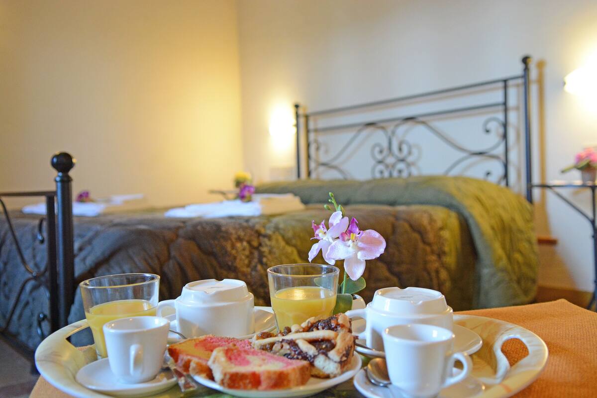 A breakfast tray with toast, fruit, and juice, set on a table in a hotel room with a bed and decorative pillows in the background.