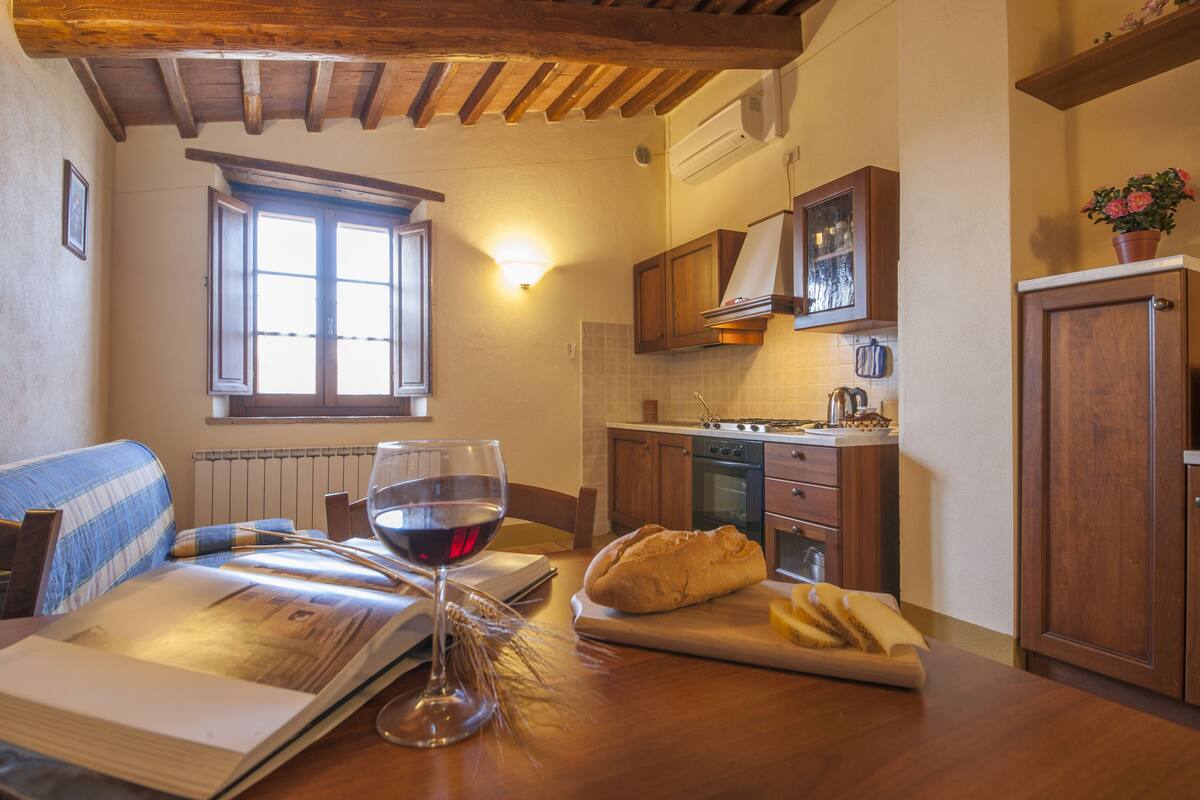 A cozy kitchen with wooden cabinets, a dining table with a book, a glass of red wine, and a loaf of bread.