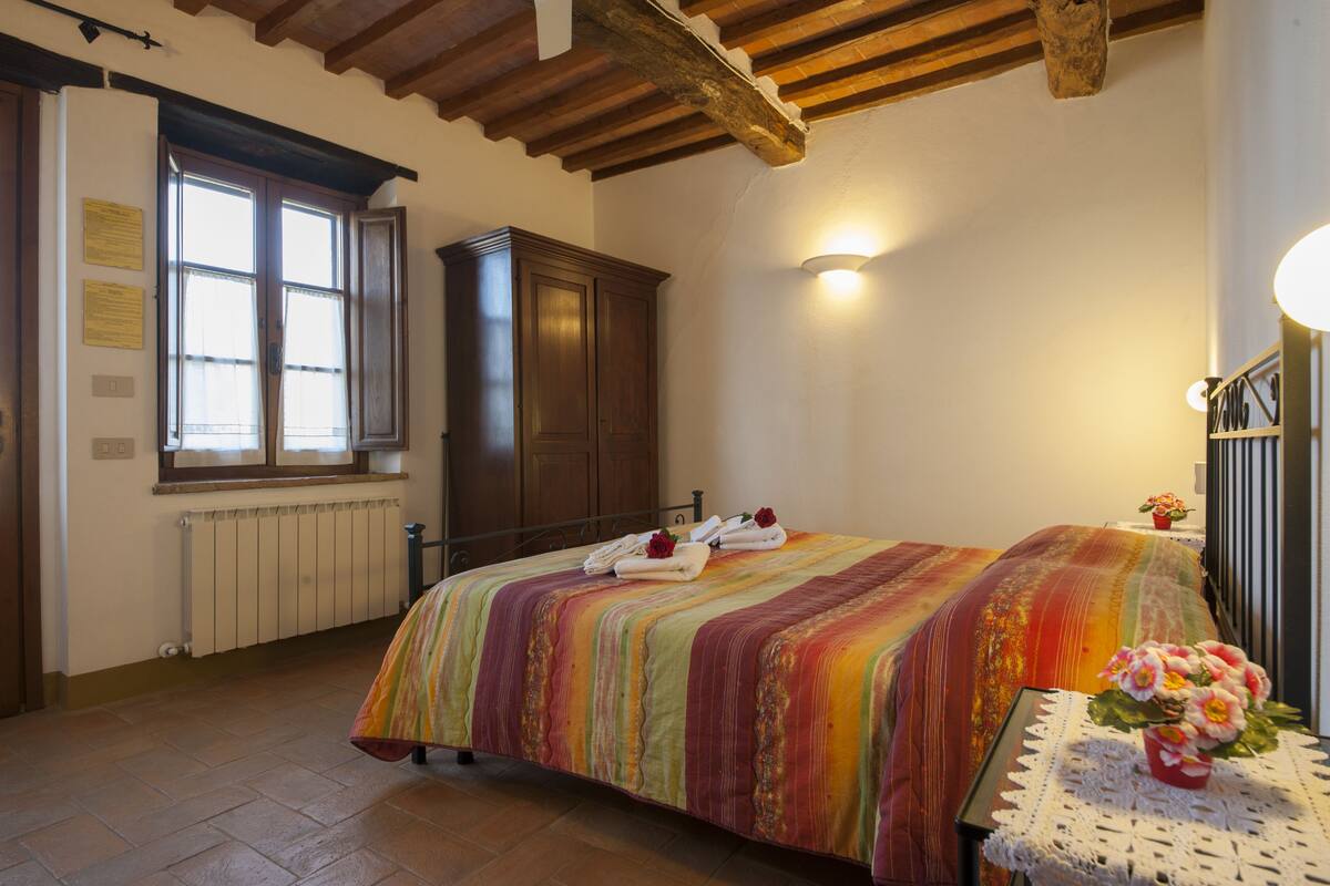 A bedroom with a bed, a wooden wardrobe, a radiator, a window, and a wall-mounted light fixture.