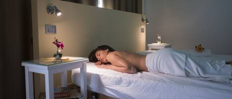 A single bed with a white mattress and a white blanket, a small bedside table with a lamp, a book, and a flower.