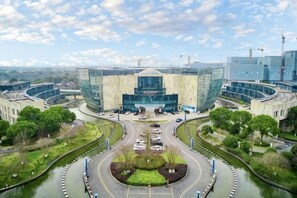 Een modern gebouw met een centrale glazen gevel, omringd door een aangelegde omgeving met een cirkelvormige oprit en een kanaal.