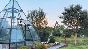 A glass greenhouse structure with a geometric design, surrounded by a garden with various plants and a wooden deck.