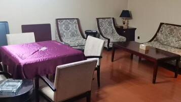 A room with a purple table, two patterned armchairs, a sofa, a small round table, and a lamp on a nightstand.