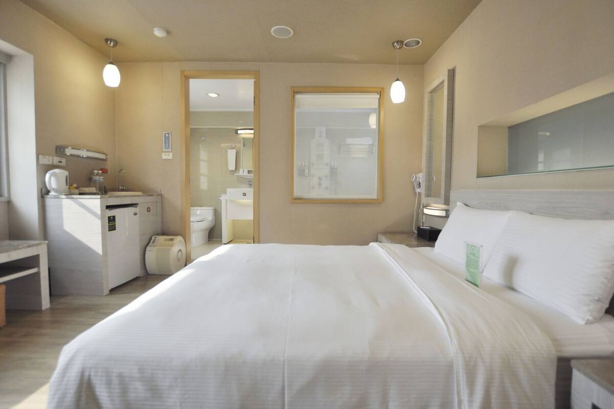 A hotel room with a bed, bedside table, a desk with a kettle, a mirror, and a view of the bathroom.