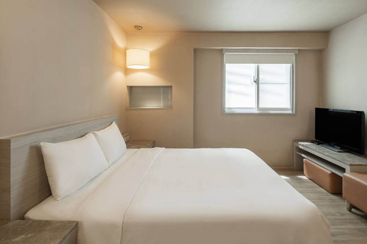 A hotel room with a large bed, a TV on a wooden stand, a window with blinds, and a bedside lamp.