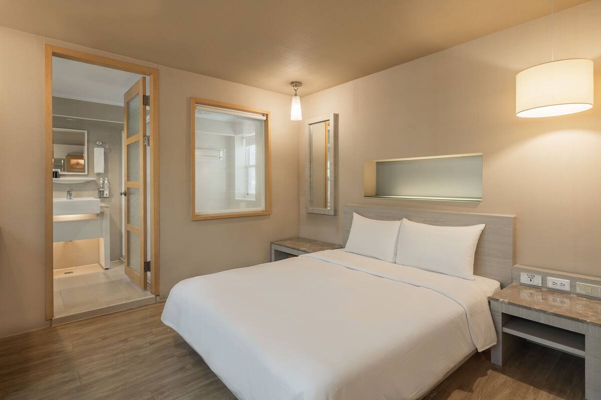 A hotel room with a bed, bedside tables, a nightstand, a mirror, and a bathroom visible through an open door.