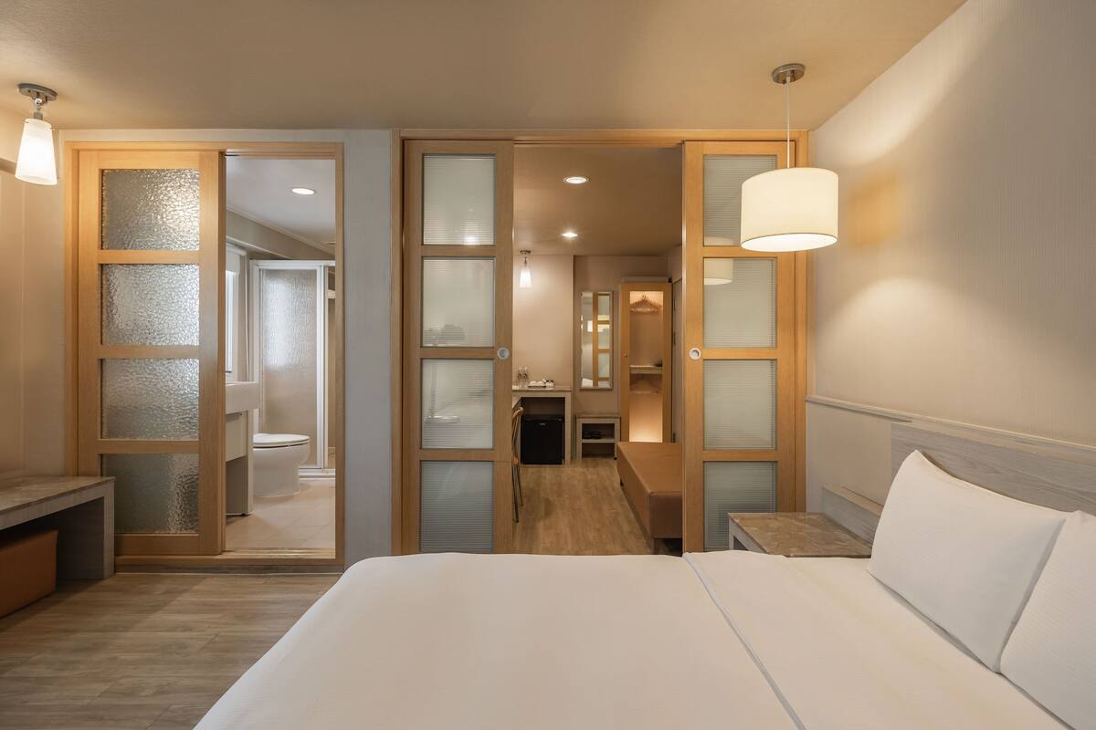 A hotel room with a large bed, a desk, a mirror, and a bathroom visible through a glass door.
