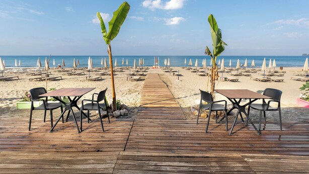 A wooden deck with tables and chairs, leading to a beach with umbrellas and lounge chairs. - Krizantem Hotel - All Inclusive (Alanya)