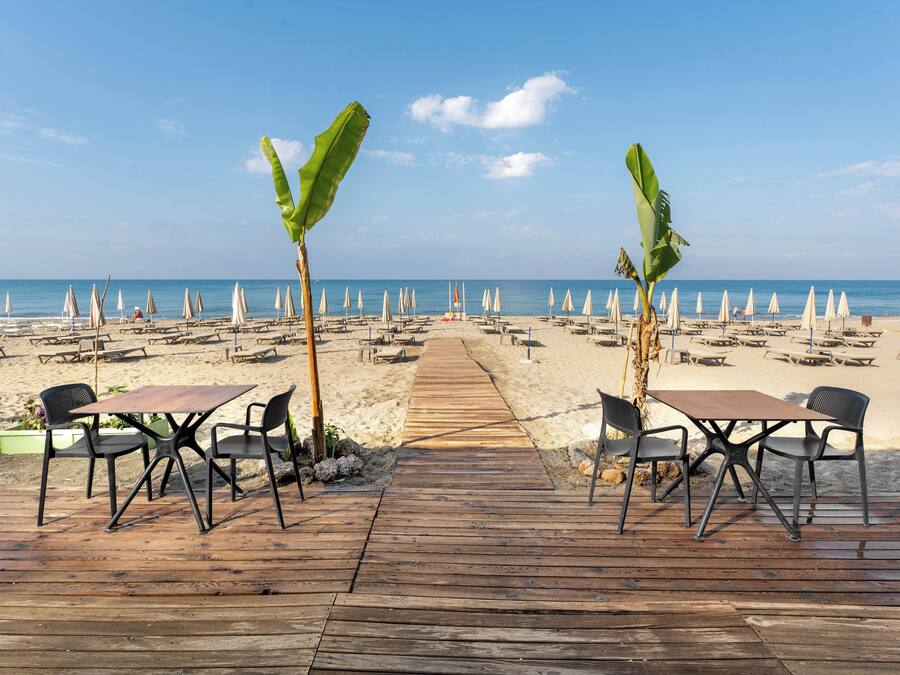 A wooden deck with tables and chairs, leading to a beach with umbrellas and lounge chairs.