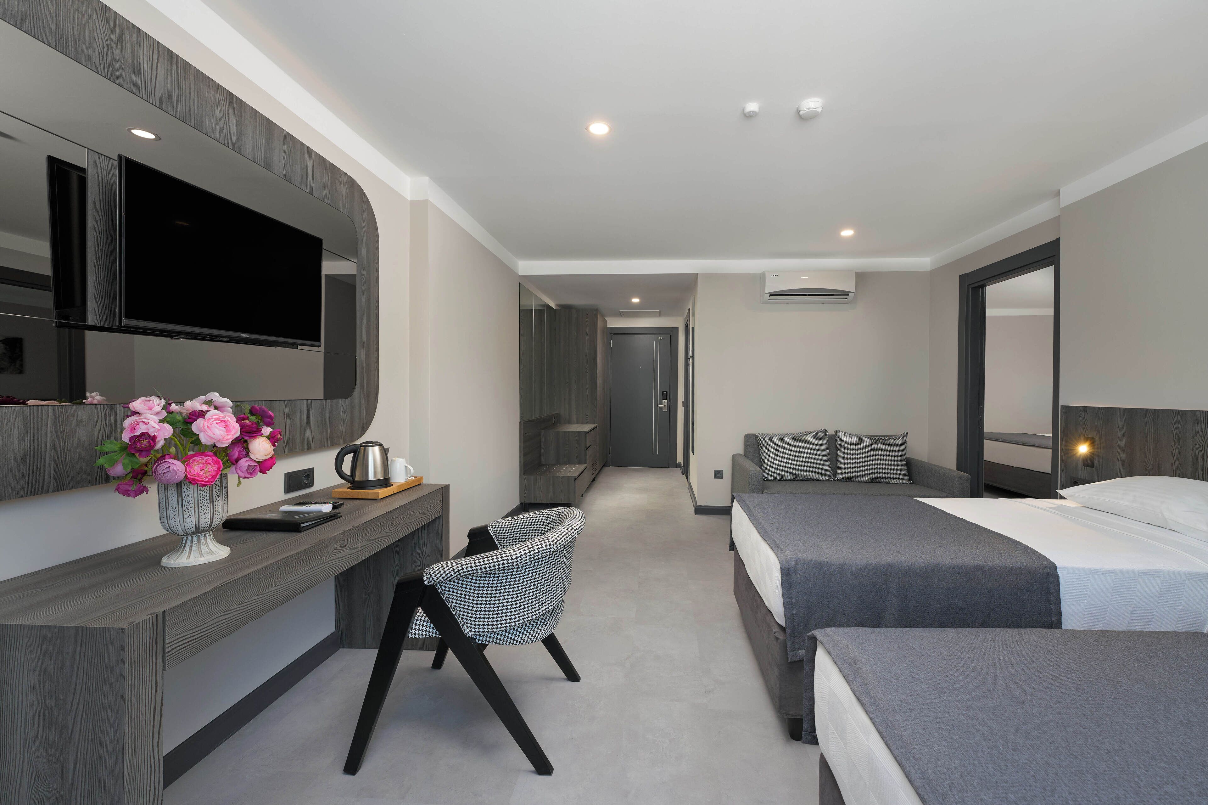 A modern hotel room with a large bed, a desk with a chair, a flat-screen TV mounted on the wall, and a small seating area with a chair and a vase of flowers.