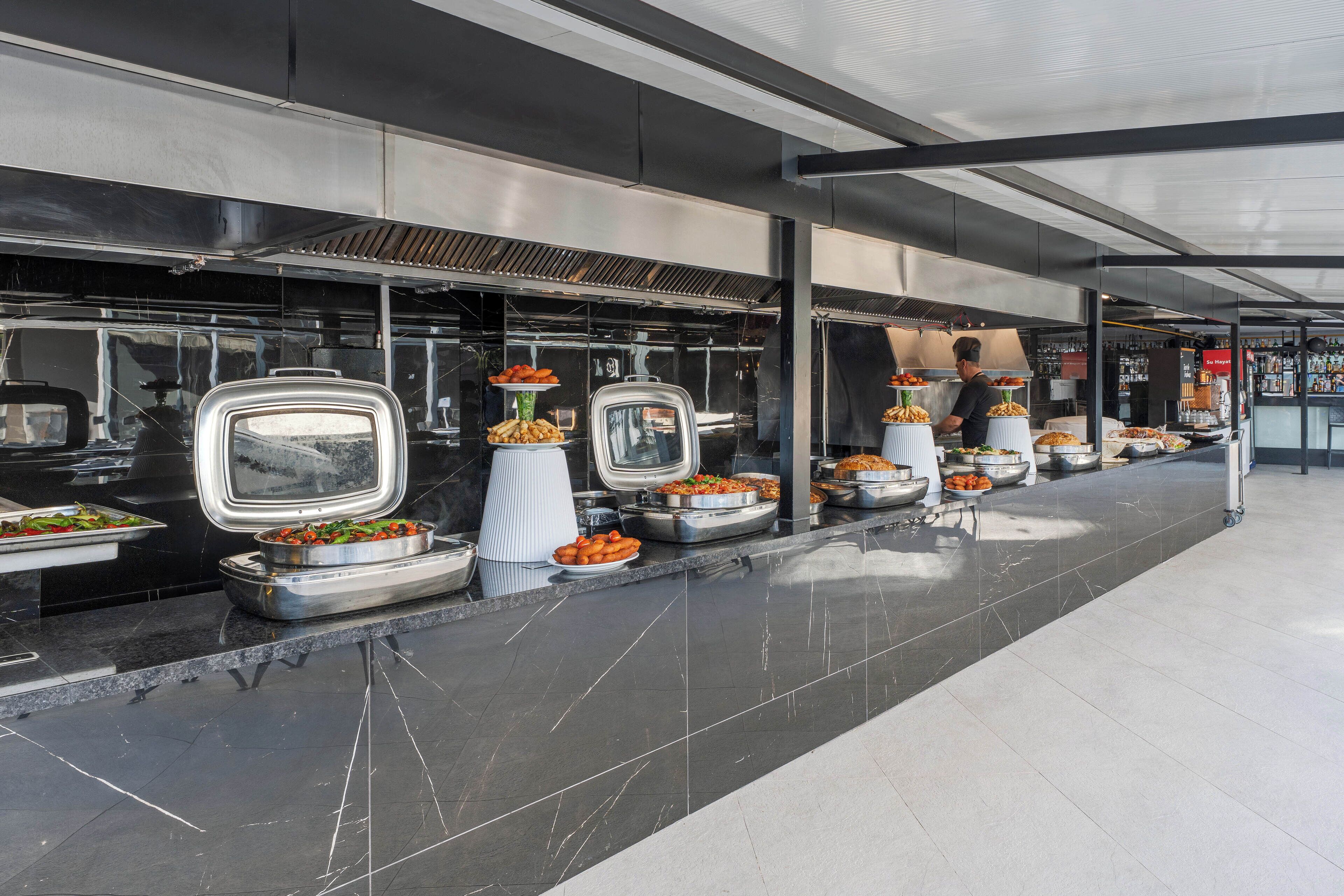 A buffet setup with various food items displayed in trays and bowls, arranged along a long counter with a tiled floor.