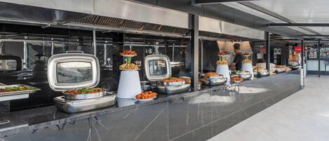 A buffet setup with various food items displayed in trays and bowls, arranged along a long counter with a tiled floor.
