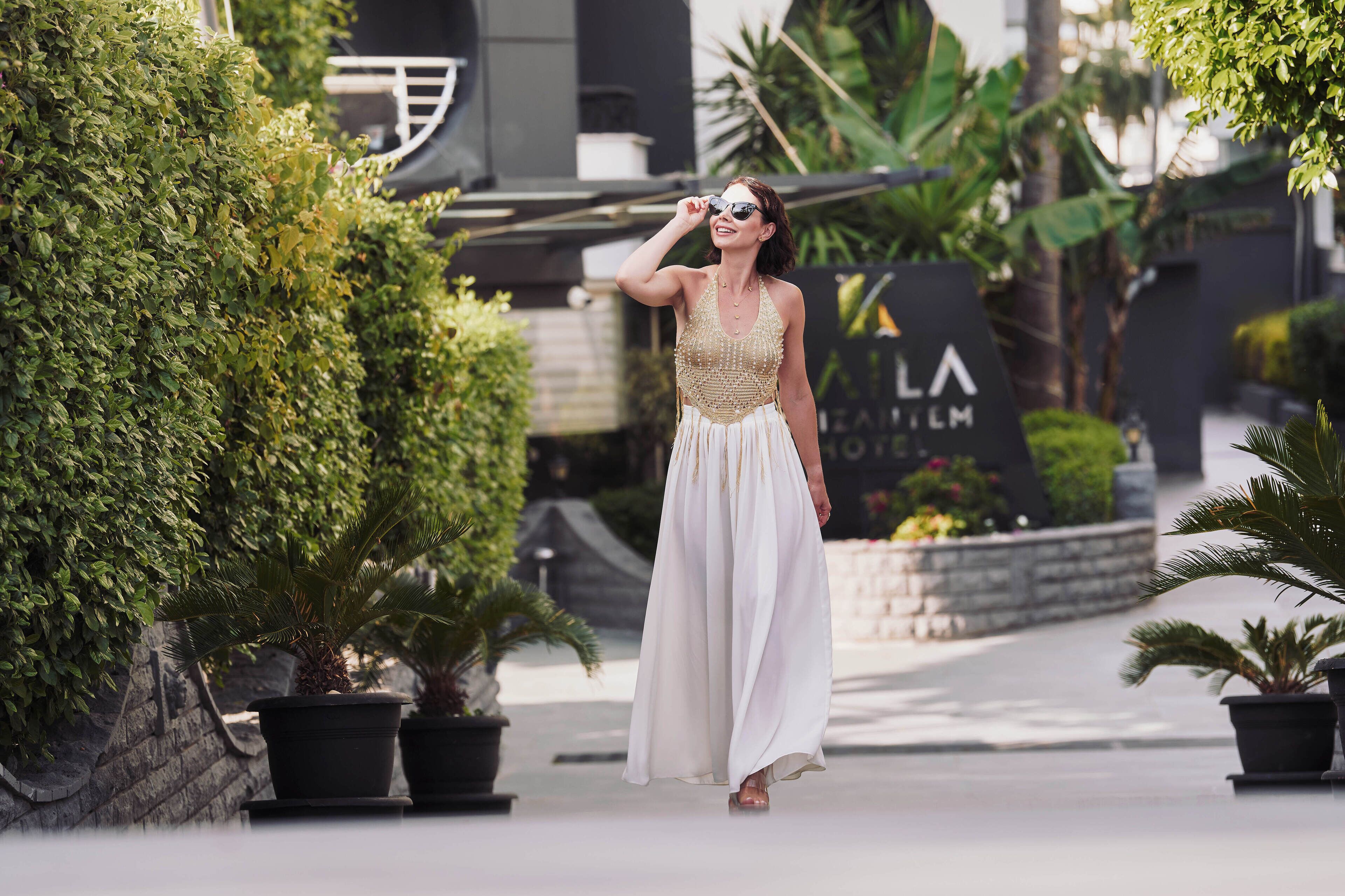 A woman in a white dress walking on a paved path with potted plants and a sign in the background.