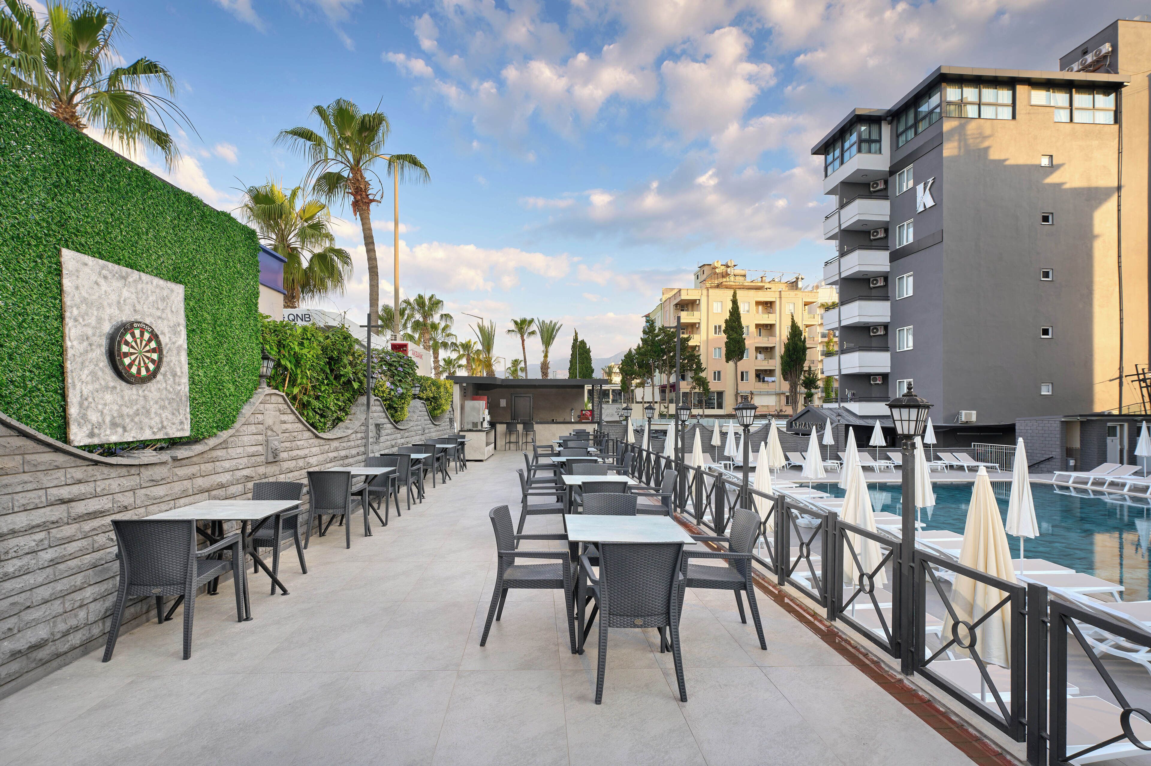 An outdoor patio with tables and chairs, a dartboard, a pool, and a building with balconies.