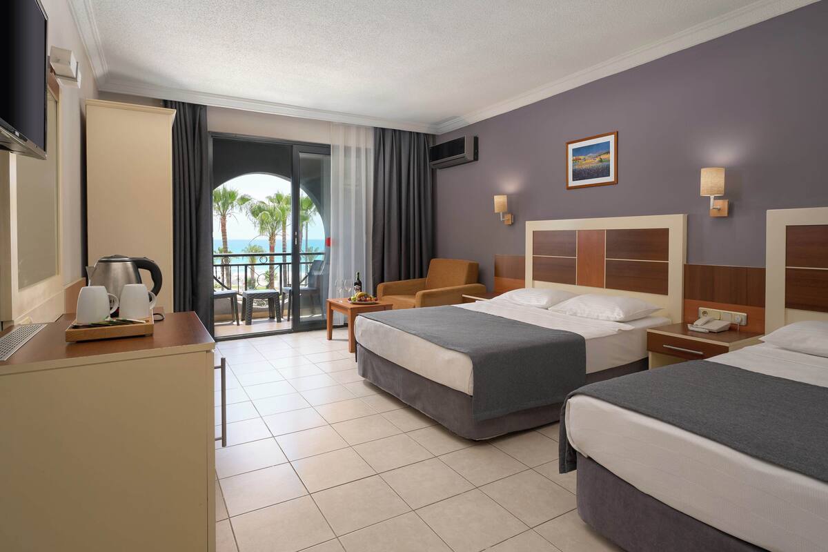 A hotel room with two beds, a desk with a kettle, a chair, a small table with fruit, and a balcony with a view of palm trees.