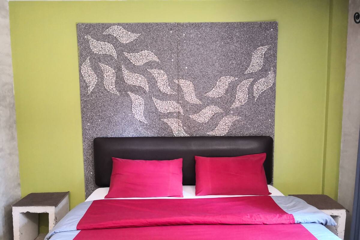 Un lit avec une couvre-lit rouge et bleue, deux oreillers rouges et une tête de lit ornée d’un motif en mosaïque.