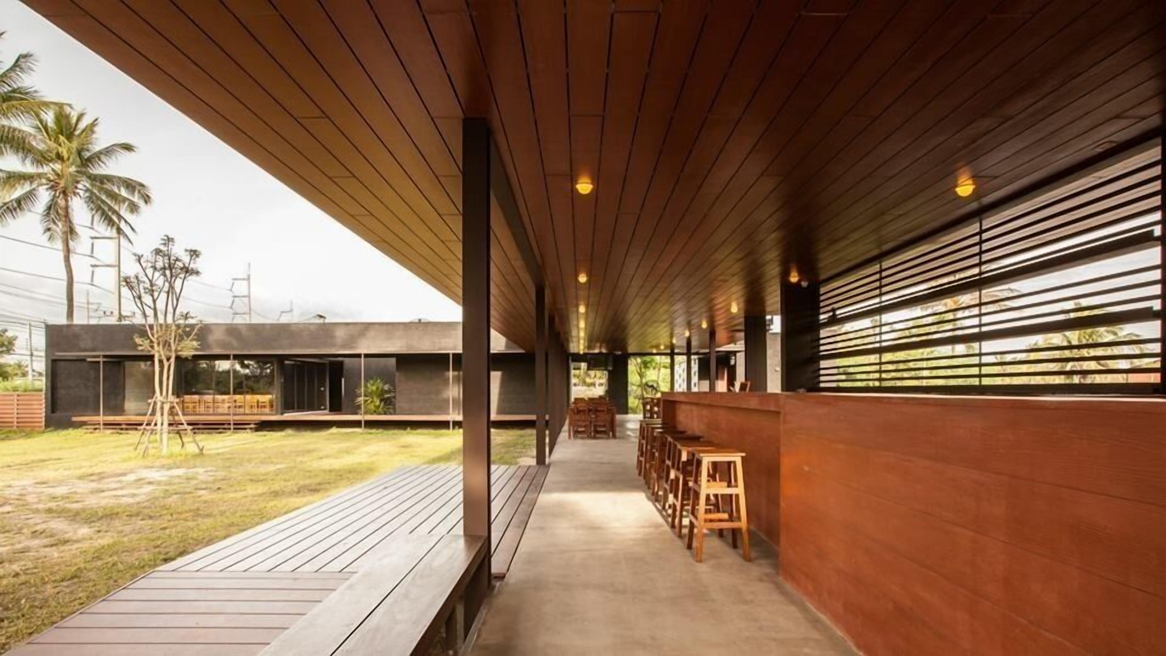 A modern outdoor patio with wooden flooring and benches, a covered seating area with a wooden ceiling, and a view of a modern building with large windows.