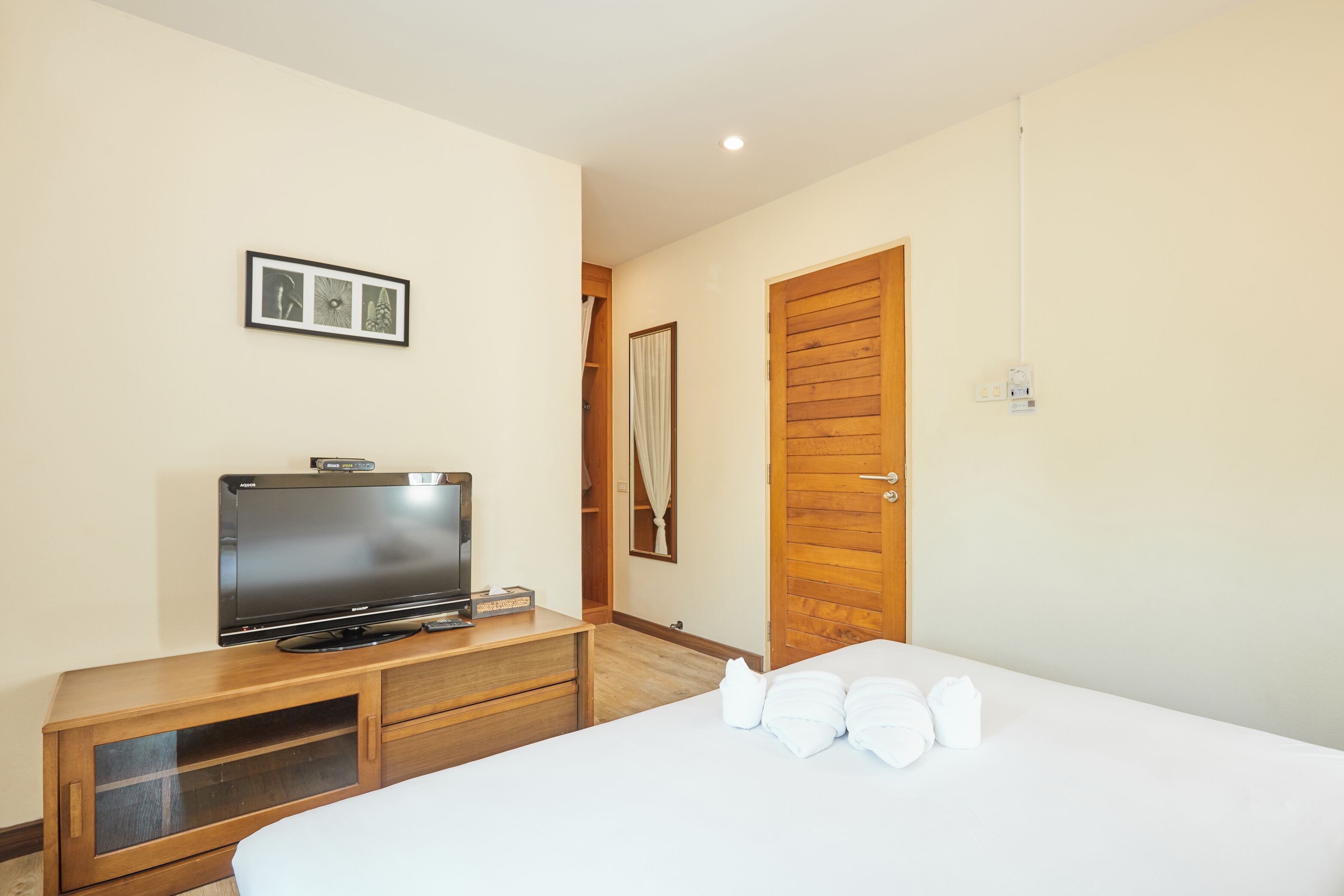 Quarto de hotel com uma cama, uma televisão sobre um móvel de madeira, uma porta de madeira e um quadro fixado na parede.