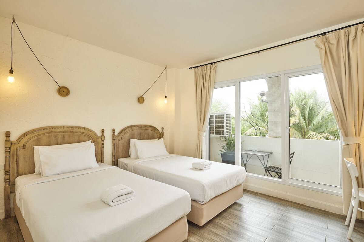 A hotel room with two beds, a balcony with a table and chairs, and a view of palm trees.