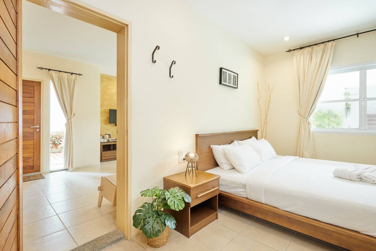 A hotel room with a wooden bed, a bedside table with a lamp, a potted plant, and a view of the outside.