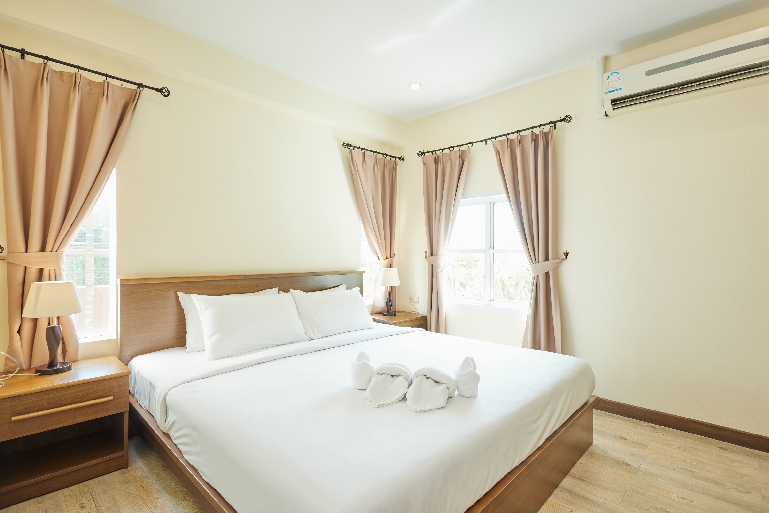 A hotel room with a large bed, two bedside tables, a nightstand, a window with curtains, and an air conditioning unit.