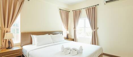 A hotel room with a large bed, two bedside tables, a nightstand, a window with curtains, and an air conditioning unit.