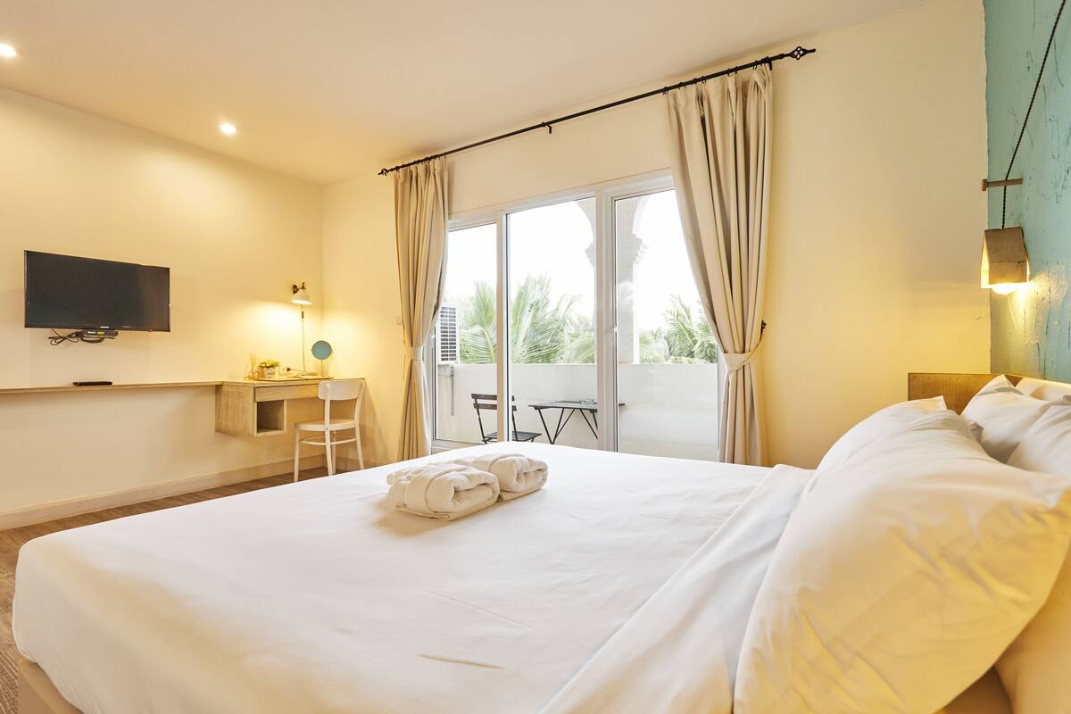 A hotel room with a bed, a desk, a chair, a TV, and a balcony with a view of palm trees.