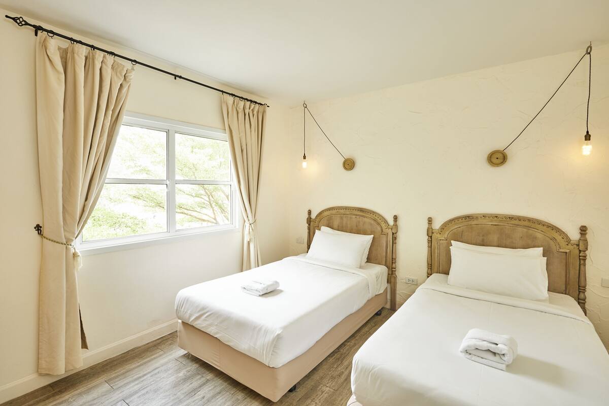 A room with two beds, a window with curtains, a wooden headboard, and a wall-mounted light fixture.