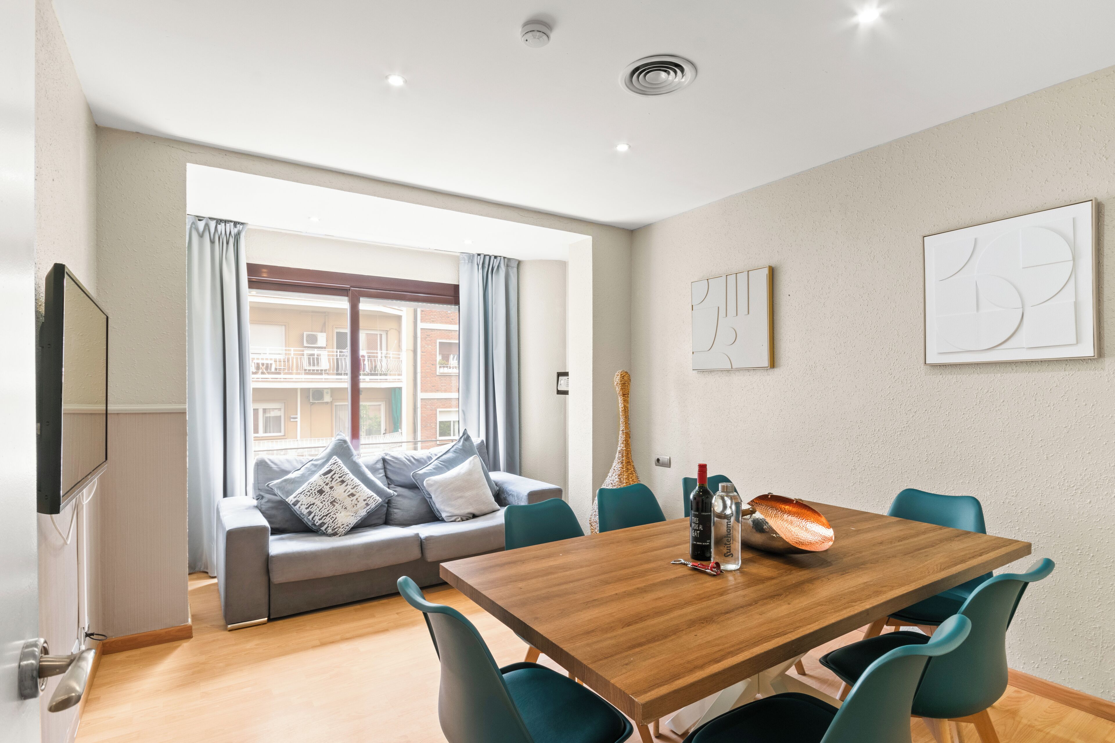 A modern living room with a wooden dining table, teal chairs, a grey sofa with patterned cushions, a flat-screen TV, and a large window with curtains.