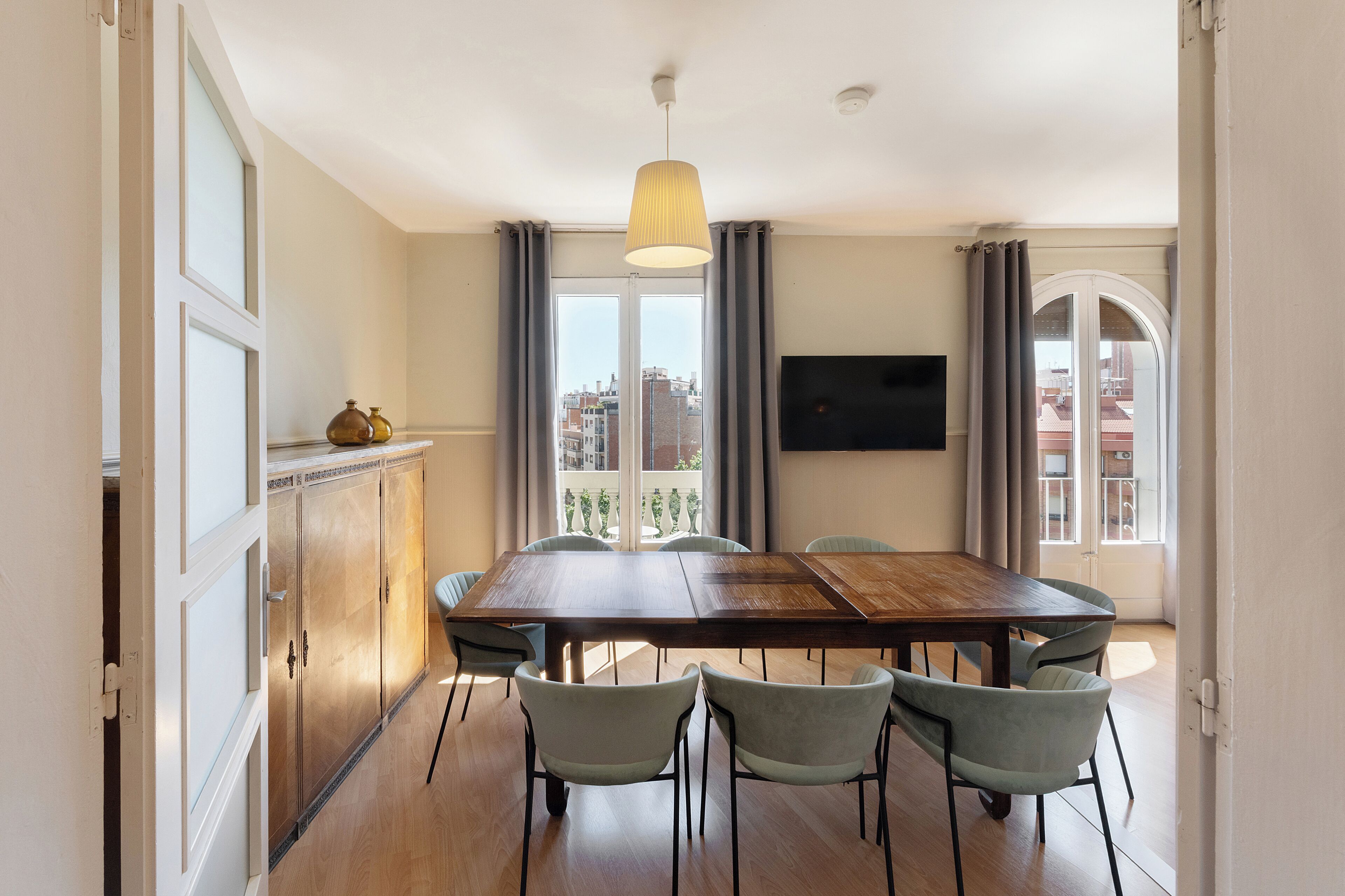 A dining area with a wooden table, four chairs, a hanging light fixture, a television on the wall, and a view of buildings through large windows.
