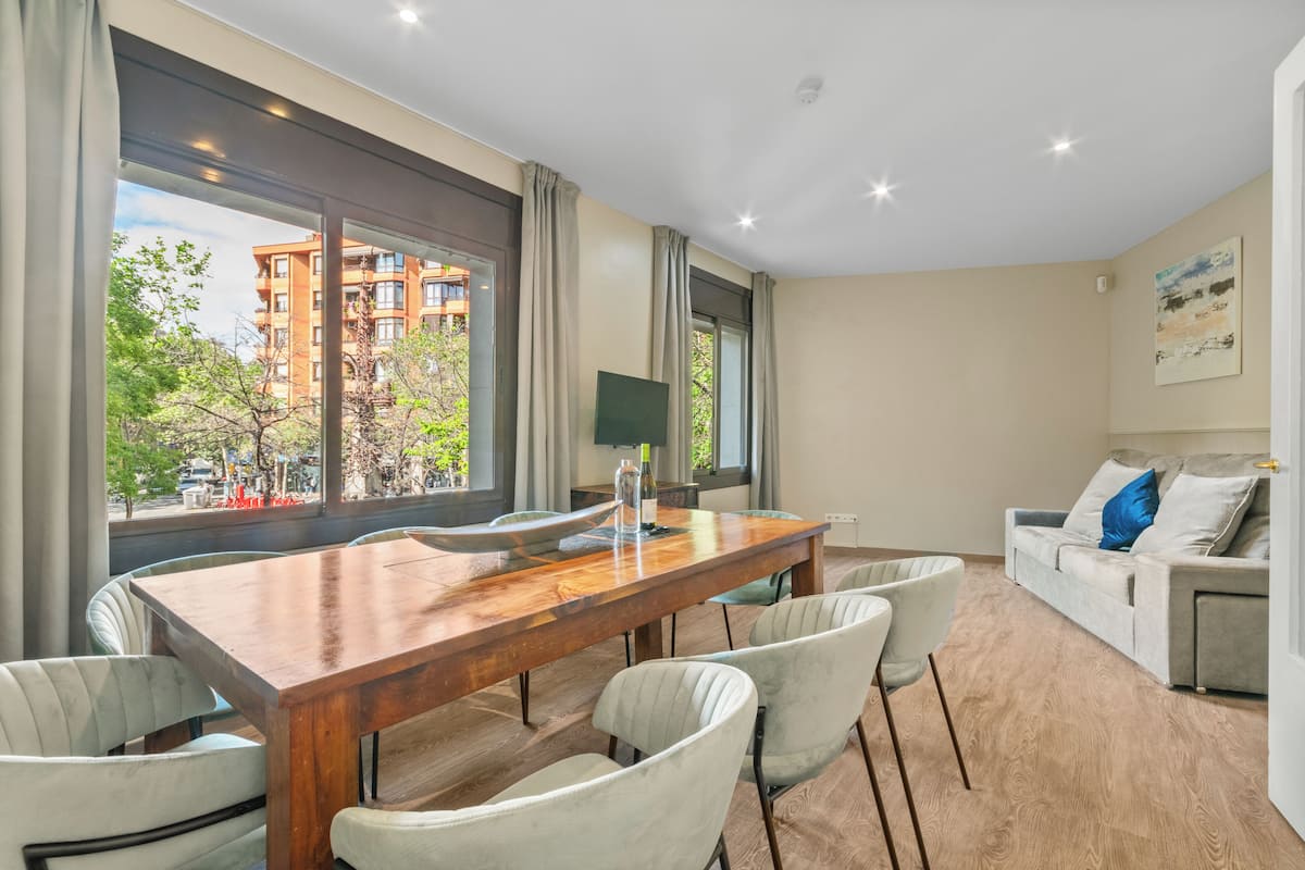 Una sala de estar moderna con una mesa de comedor de madera, sillas de color claro, un sofá y un televisor.