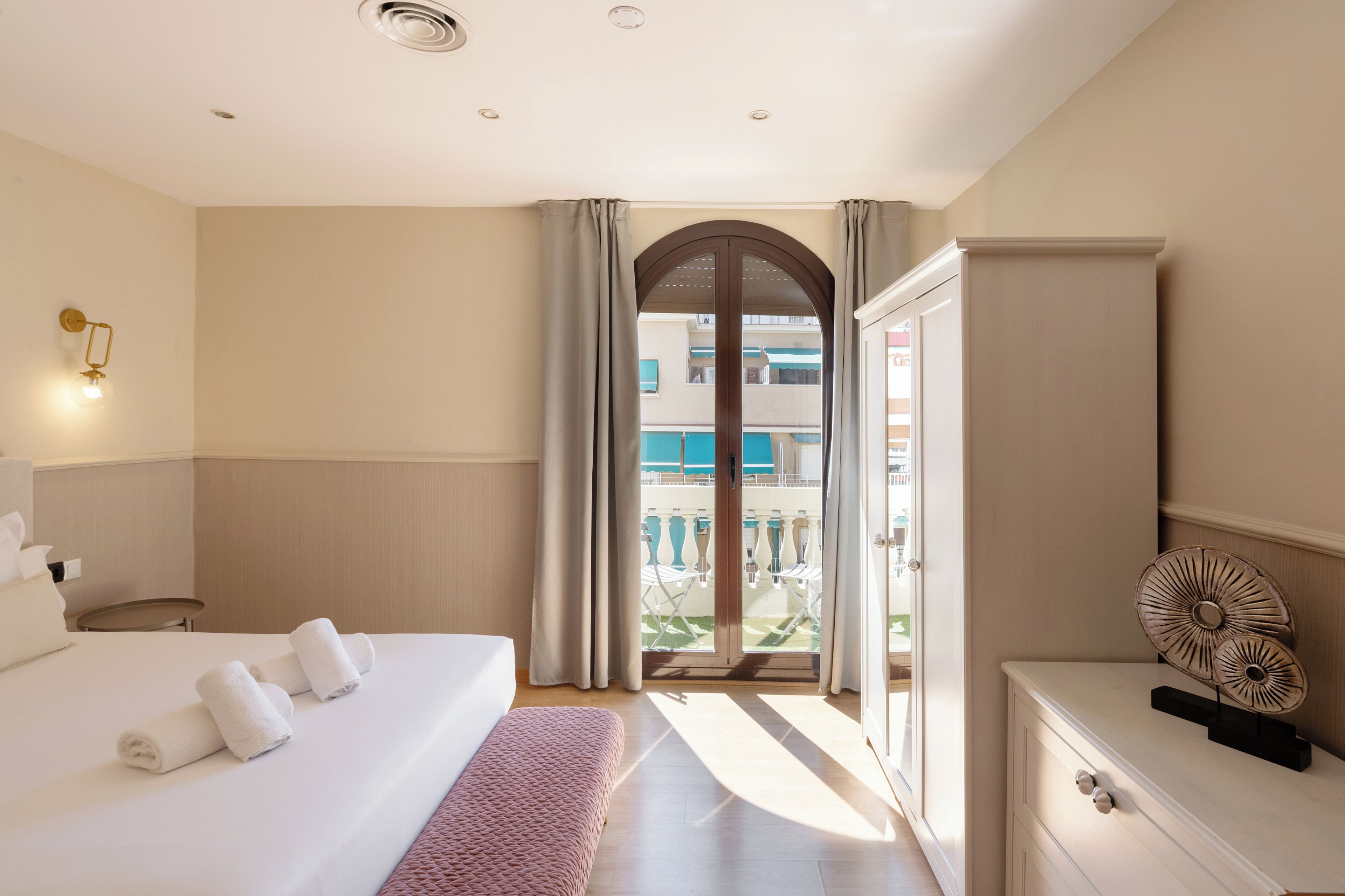 A hotel room with a bed, a bedside table, a wardrobe, a window with curtains, and a view of the outside balcony.