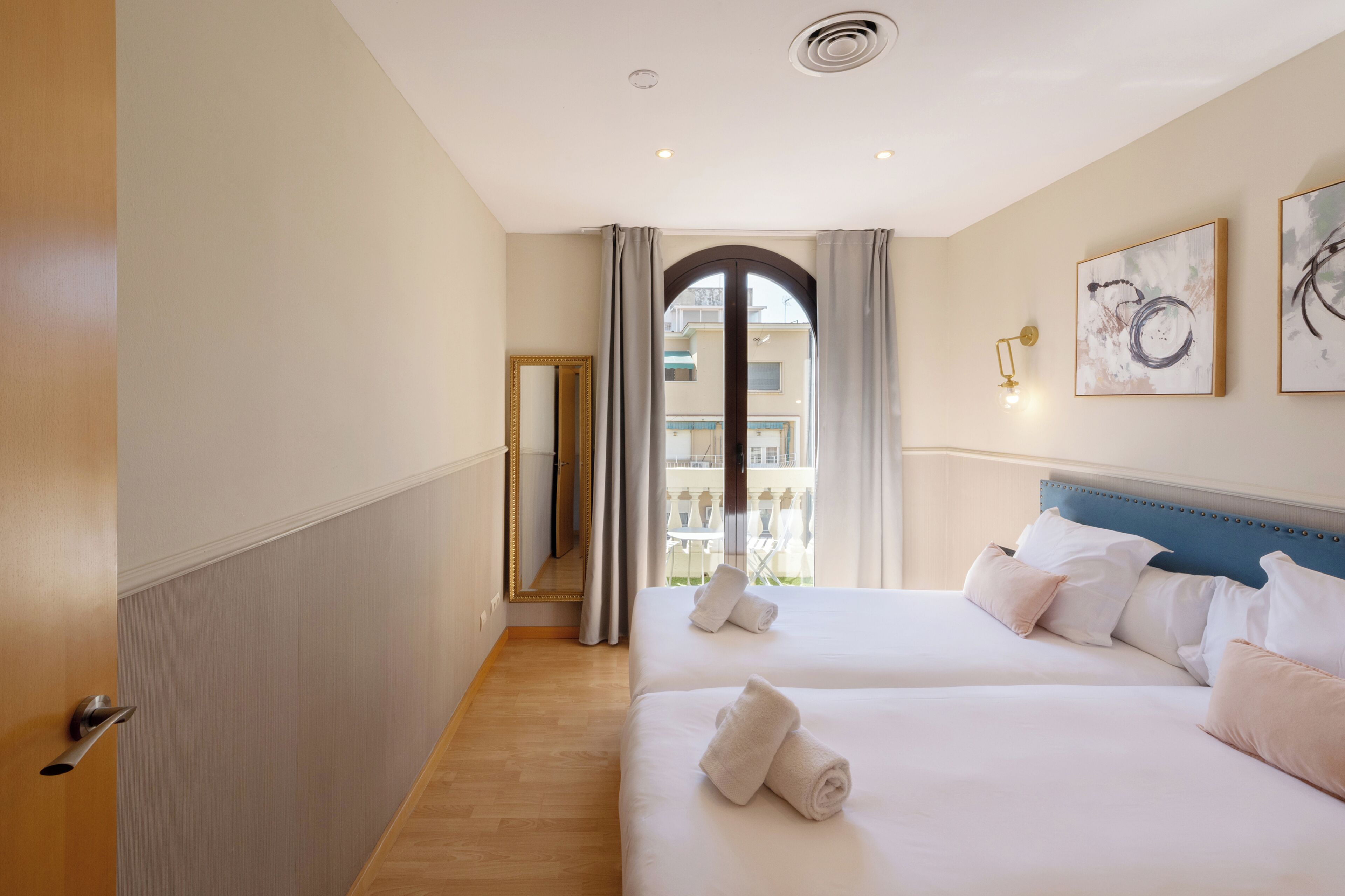 A hotel room with two beds, a large mirror, a view through a window, and artwork on the wall.