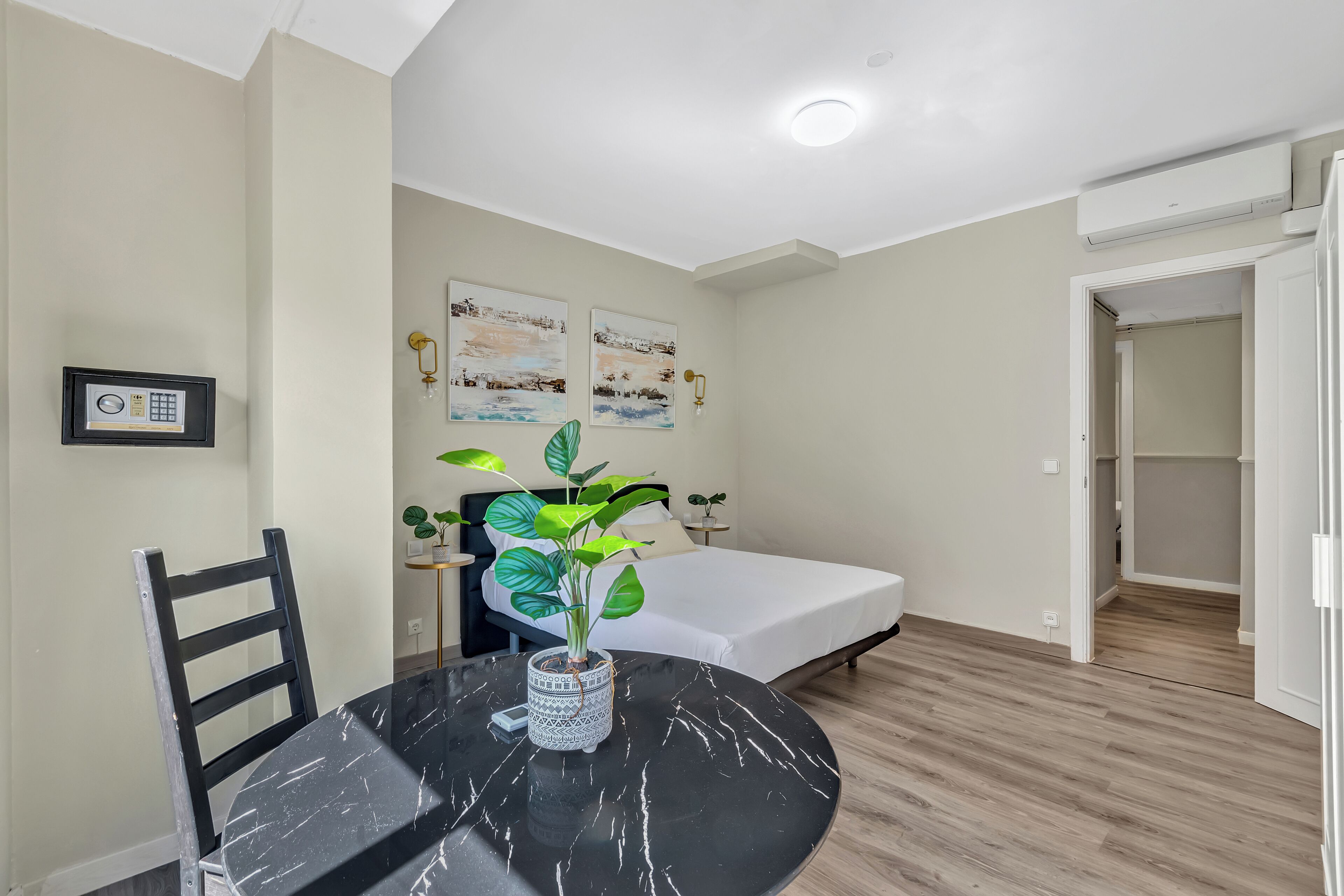 A compact room with a bed, a dining table with chairs, a potted plant, and two wall-mounted paintings.