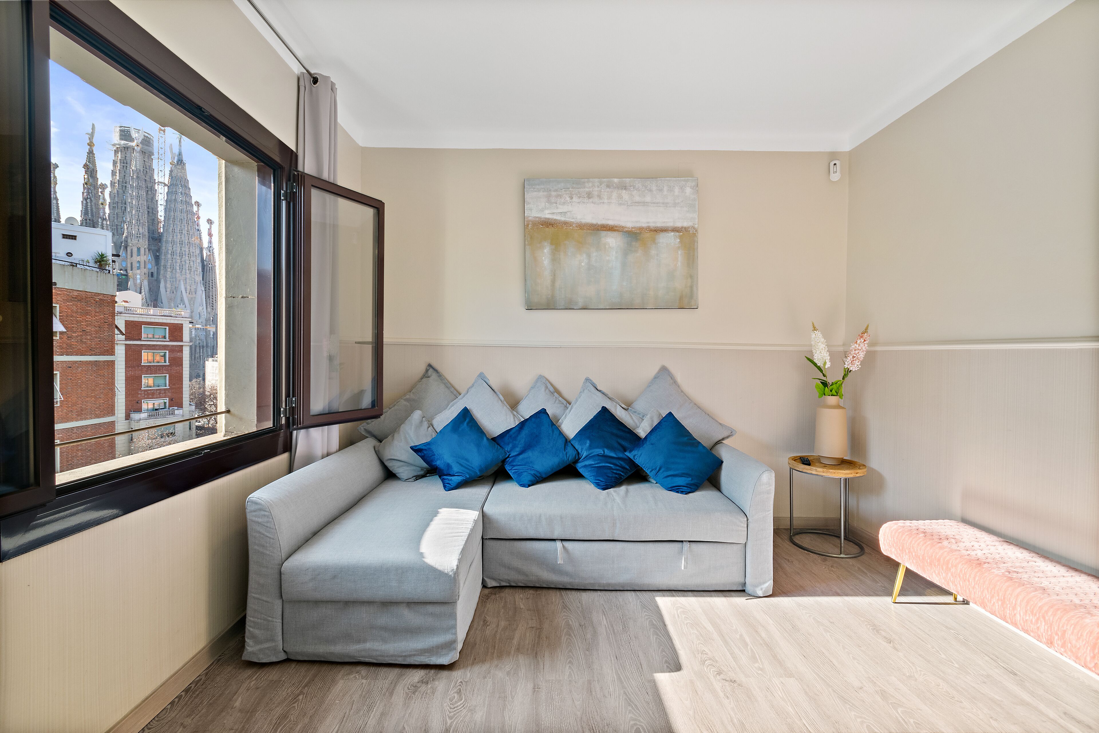 Ein modernes Wohnzimmer mit einem grauen Ecksofa, blauen Kissen, einem kleinen runden Tisch und einer pinkfarbenen Bank.