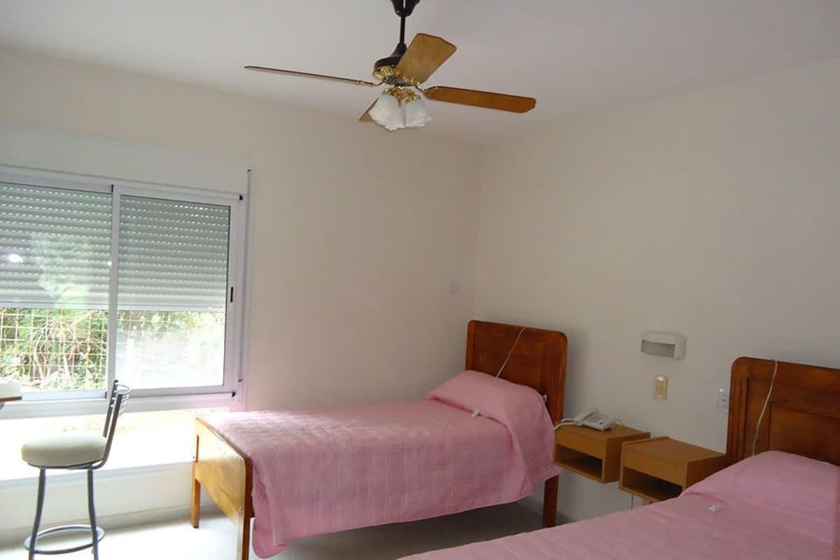 Un dormitorio con dos camas individuales, un ventilador de techo, una mesita de noche con teléfono y una silla cerca de una ventana con persianas.