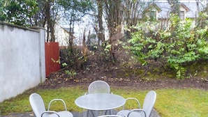 Un patio con cuatro sillas de metal y una mesa, rodeado de césped y un muro de piedra bajo.