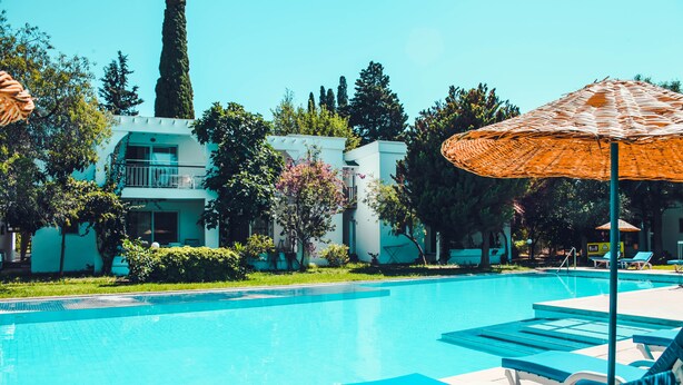 Una piscina con sillones de descanso y sombrillas, rodeada de vegetación y edificios modernos.